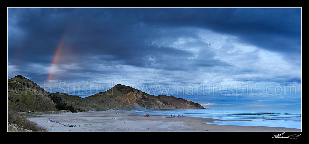 Image of Kaiaua Bay Beach moody twilight panorama with rainbow as rain starts and storm clouds gather, with Marau Point centre. People retrieving or launching small boat, Tolaga Bay, Gisborne District, Gisborne Region, New Zealand (NZ) stock photo image