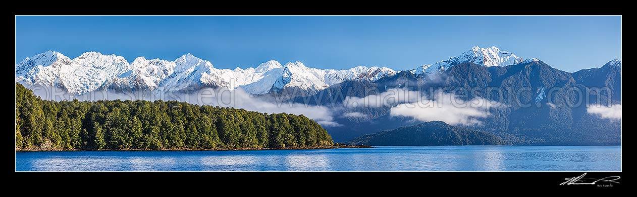 Image of Lake Manapouri morning winter calm with Stony Point bushy headland in front of Kepler Moutains under winter snow. Rona Island right. Panorama, Te Anau, Fiordland National Park, Southland District, Southland Region, New Zealand (NZ) stock photo image