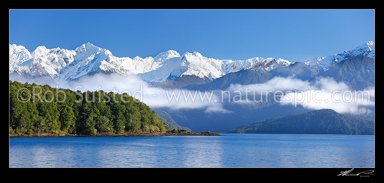 Image of Lake Manapouri morning winter calm with Stony Point bushy headland in front of Kepler Moutains under winter snow. Rona Island right. Panorama, Te Anau, Fiordland National Park, Southland District, Southland Region, New Zealand (NZ) stock photo image