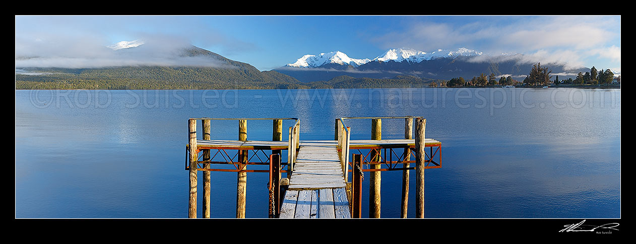 Image of  Lake Te Anau jetty on a calm frosty morning in winter with the Murchison Mountains behind in snow. Te Anau township at right. Panorama, Lake Te Anau, Fiordland National Park, Southland District, Southland Region, New Zealand (NZ) stock photo image