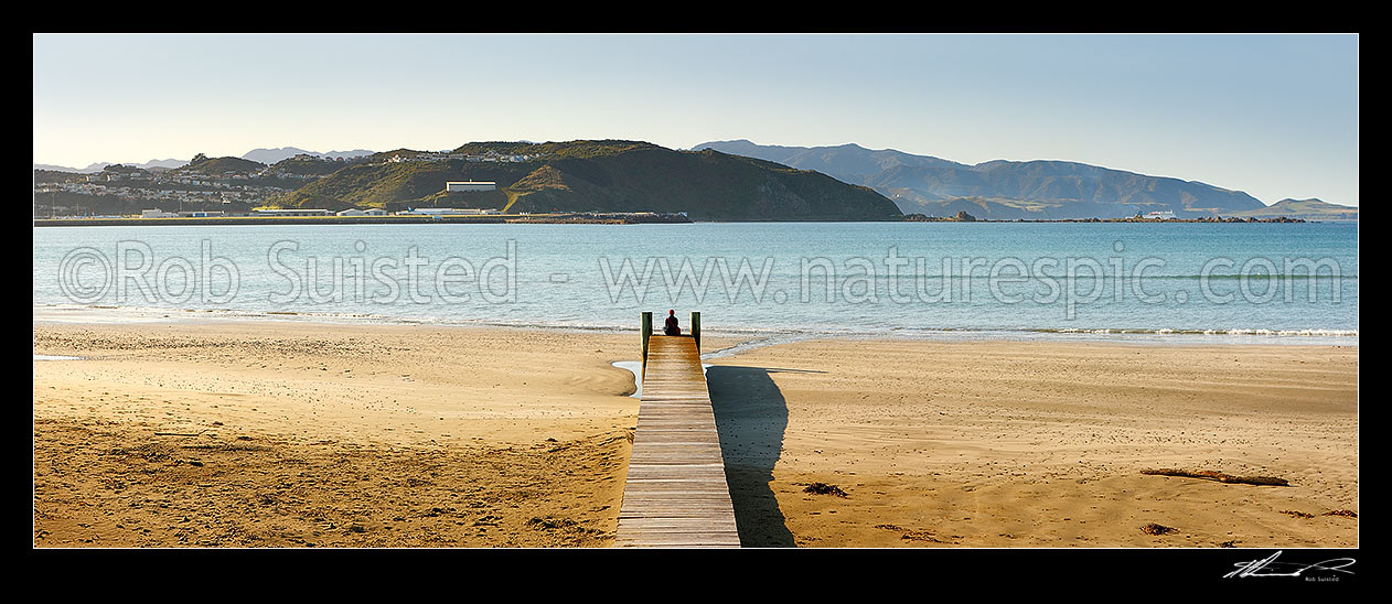 Image of Lyall Bay Beach with person sitting or meditating on end of wooden boardwalk on a fine spring morning, with Wellington Airport beyond. Panorama, Lyall Bay, Wellington, Wellington City District, Wellington Region, New Zealand (NZ) stock photo image