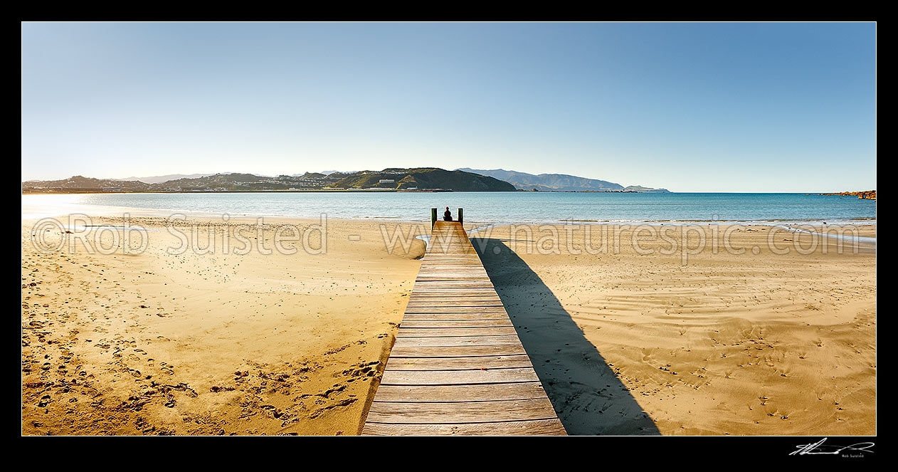 Image of Lyall Bay Beach with person sitting or meditating on end of wooden boardwalk on a fine spring morning, with Wellington Airport beyond. Panorama, Lyall Bay, Wellington, Wellington City District, Wellington Region, New Zealand (NZ) stock photo image