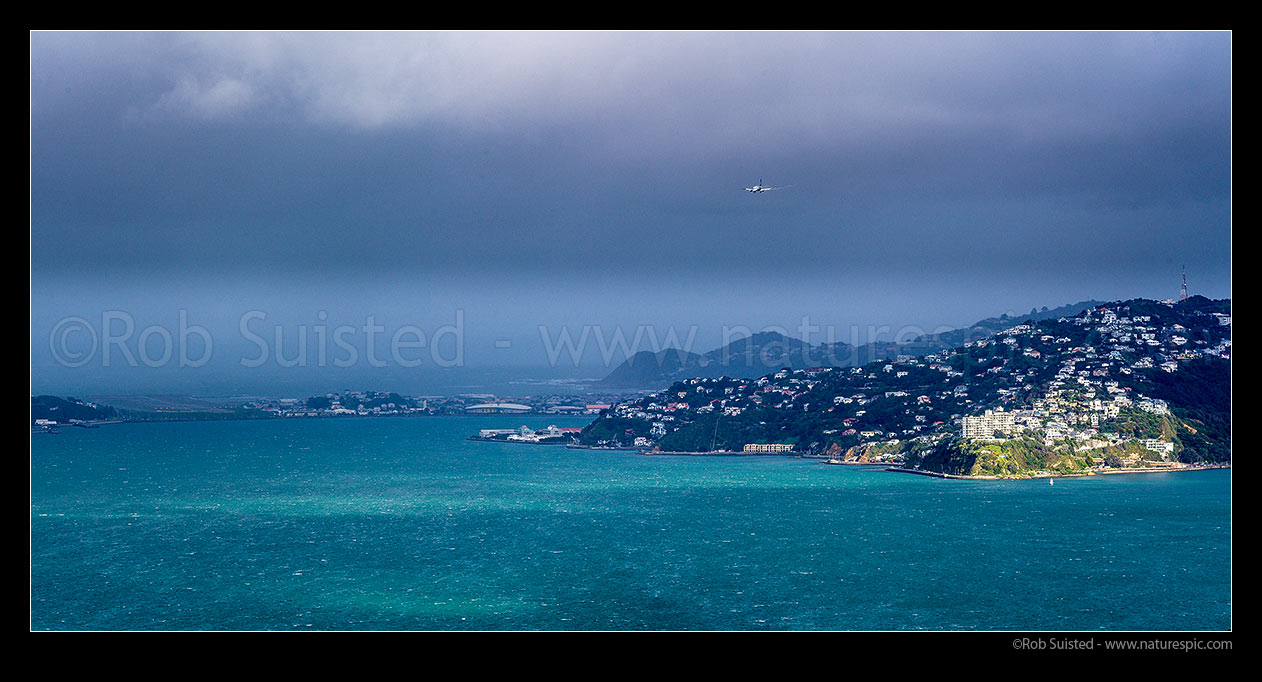 Image of Wellington airport with Air New Zealand airplane landing into a biting southerly storm front gale, with last sun rays on Point Jerningham. Panorama, Kilbirnie, Wellington City District, Wellington Region, New Zealand (NZ) stock photo image