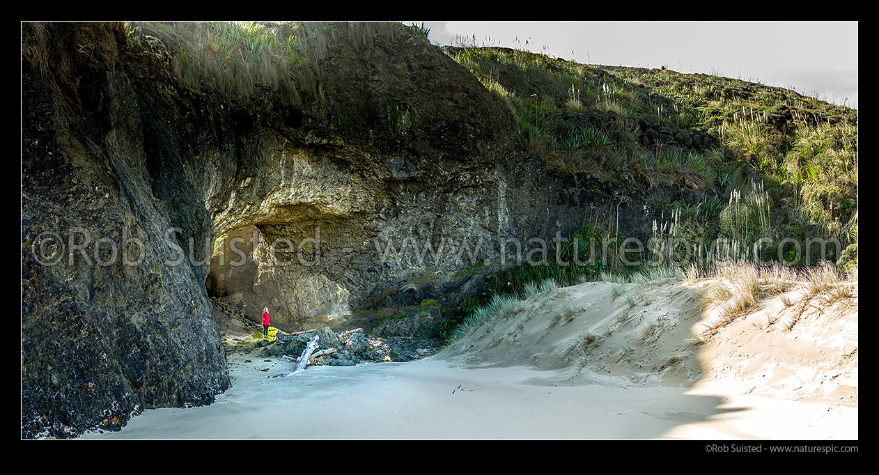 Image of Old sea cave on Mitimiti Beach, with visitor checking out the natural archway. Okaihau Pa and Moerewa Point. Panorama, Mitimiti, Far North District, Northland Region, New Zealand (NZ) stock photo image