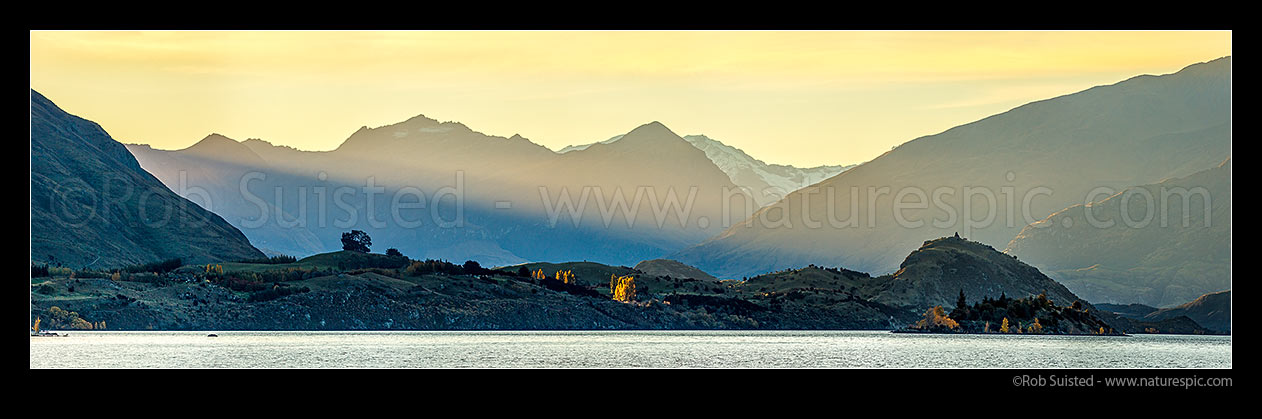 Image of Lake Wanaka, with last rays of sunlight raking across Sharks Tooth Peak, Fog Peak (2240m) and Niger Peak (centre). Matukituki Valley and Mt Aspiring National Park beyond. Autumn colours. Panorama, Wanaka, Queenstown Lakes District, Otago Region, New Zealand (NZ) stock photo image