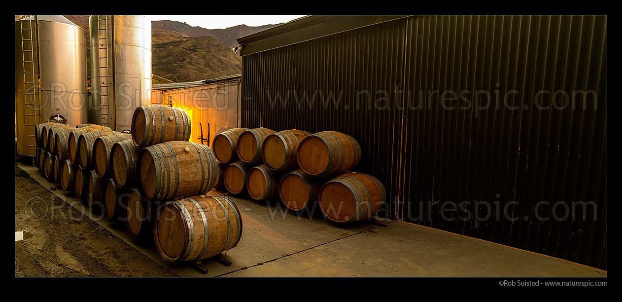 Image of Industrial commercial wine background with stainless steel tanks, vats and oak barrels stacked by warehouse at dusk. Panorama file, New Zealand (NZ) stock photo image