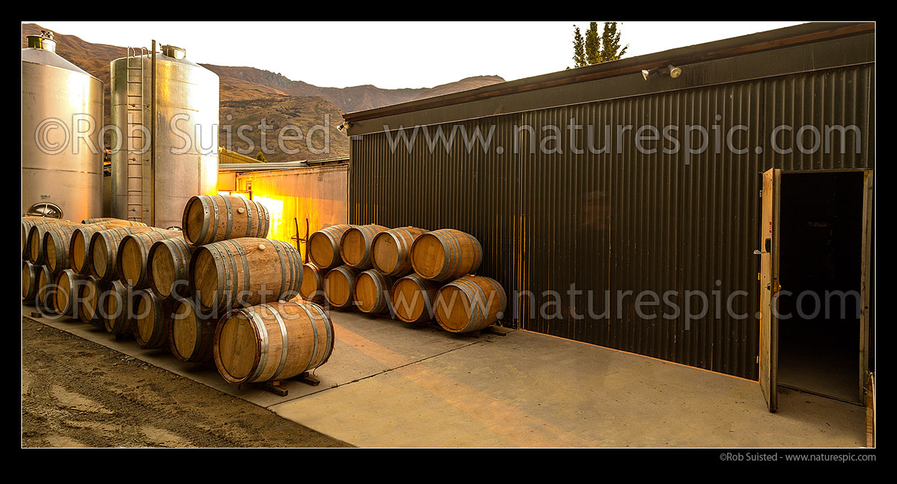 Image of Industrial commercial wine background with stainless steel tanks, vats and oak barrels stacked by warehouse at dusk. Panorama file, New Zealand (NZ) stock photo image