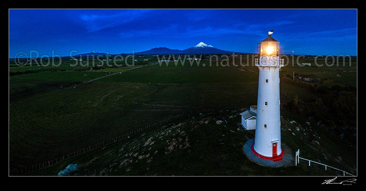 Image of Cape Egmont Lighthouse glowing at dusk, with Mt Taranaki and Egmont National Park beyond over farmland. Aerial panorama, Cape Egmont, South Taranaki District, Taranaki Region, New Zealand (NZ) stock photo image