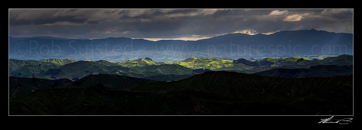 Image of Wairarapa hill country moody day, with breaking sunlight lighting hills and ridges near Eketahuna. Panorama view with northern Tararua Ranges behind. Northern Wairarapa, Pori, Tararua District, Manawatu-Wanganui Region, New Zealand (NZ) stock photo image