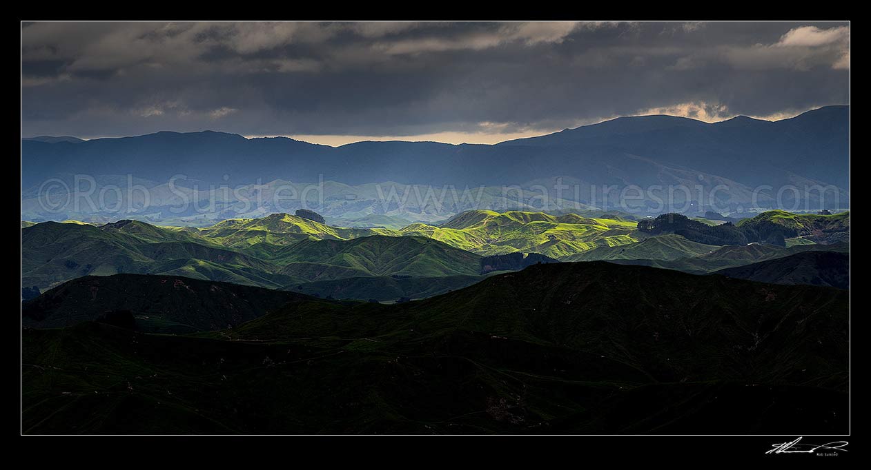 Image of Wairarapa hill country moody day, with breaking sunlight lighting hills and ridges near Eketahuna. Panorama view with northern Tararua Ranges behind. Northern Wairarapa, Pori, Tararua District, Manawatu-Wanganui Region, New Zealand (NZ) stock photo image