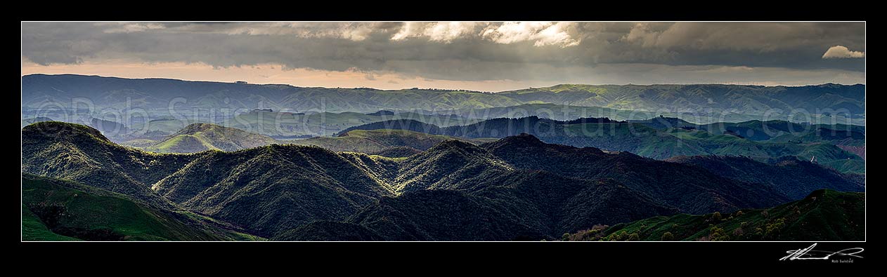 Image of Wairarapa hill country moody day, with breaking sunlight lighting hills and ridges near Eketahuna. Panorama view with northern Tararua Ranges behind. Northern Wairarapa, Pori, Tararua District, Manawatu-Wanganui Region, New Zealand (NZ) stock photo image